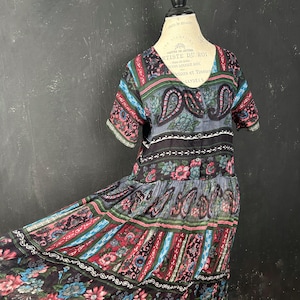 May include: A colorful, patterned dress with short sleeves and a V-neck. The dress features a mix of floral, paisley, and striped designs in shades of red, blue, green, and black. The dress is displayed on a mannequin.