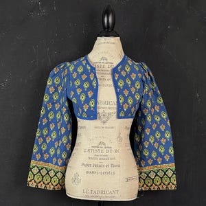 Peut inclure: Une veste courte bleue ornée d'un motif répétitif de formes en goutte d'eau vertes et orange. Le vêtement présente de longues manches larges avec une bordure décorative noire, verte et orange aux poignets et un liseré bleu sur le devant.