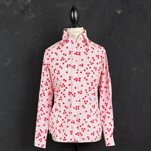 May include: A long-sleeve button-down shirt featuring a white base with a repeating pattern of small red circular dots. The shirt has a pointed collar, a front button placket, and long sleeves with buttoned cuffs, displayed on a mannequin torso.