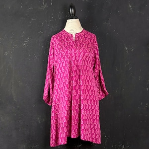 Op de afbeelding: Een tuniek met lange mouwen in een levendige magentaroze kleur, voorzien van een herhalend druppelvormig paisley-patroon in een lichtere roze tint. Het kledingstuk heeft een split-V-hals met subtiele verticale plooien vanaf de kraag.
