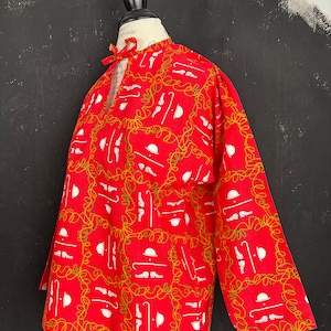 Può includere: Una camicia rossa a maniche lunghe con un motivo grafico bianco ripetuto di bombette, bastoni da passeggio e scarpe, racchiuso in una griglia di linee verde lime intrecciate. La camicia presenta uno scollo rotondo con un piccolo laccio rosso.