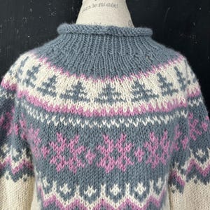May include: A grey, white, and pink knitted jumper with a high neck. The jumper features a detailed pattern of geometric and floral designs. The colour palette includes shades of grey, white, and pink, creating a cosy and textured look. The jumper is a classic piece of knitwear.