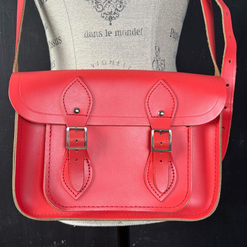 May include: A bright red satchel bag with a flap closure and two silver buckles. The bag has a shoulder strap and is made of a smooth, possibly leather, material. The bag is displayed on a mannequin.