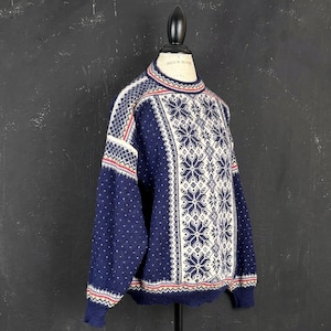 Peut inclure: Un pull en tricot à manches longues avec une base bleu marine profond ornée d'un motif central blanc de flocons de neige et de losanges. Les manches et les côtés présentent une texture à pois blancs, rehaussée de fines rayures horizontales rouges et blanches.