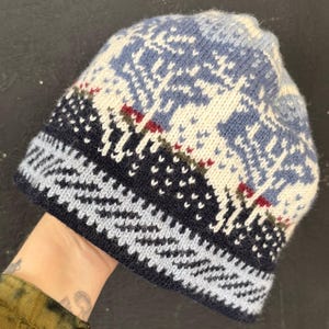 Puede incluir: Un gorro de punto con un patrón de paisaje invernal que muestra ciervos blancos, árboles y copos de nieve sobre un fondo azul y azul marino oscuro. La sección inferior presenta un borde en zigzag azul claro y marino con textura suave.