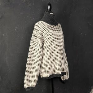 May include: An oversized sweater with a high, dark grey turtleneck. The sweater has a cream-coloured base with a repeating pattern of small, dark grey squares. The cuffs and hem are also dark grey. The sweater is displayed on a black mannequin.