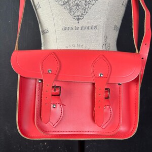 May include: A bright red satchel bag with a flap closure and adjustable shoulder strap. The bag features two buckle closures and a stitched design on the front. The bag is displayed on a mannequin.