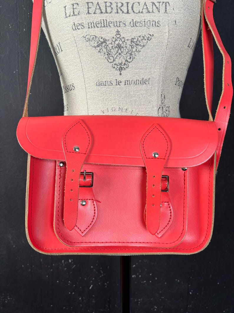 May include: A bright red satchel bag with a flap closure and adjustable shoulder strap. The bag features two buckle closures and a stitched design on the front. The bag is displayed on a mannequin.