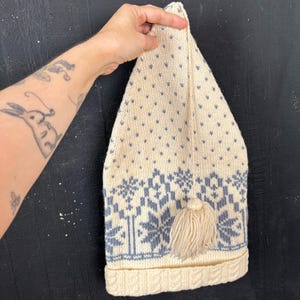 Op de afbeelding: Crèmekleurige gebreide muts met een puntige top en een kwast. De muts heeft een patroon van blauwe sneeuwvlokken en kleine hartjes. De onderrand heeft een kabelgebreid ontwerp.