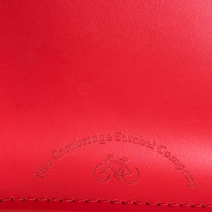 May include: Close-up of a bright red leather satchel. The Cambridge Satchel Company logo is embossed on the leather, featuring a bicycle icon. The leather has a textured appearance and a visible seam.