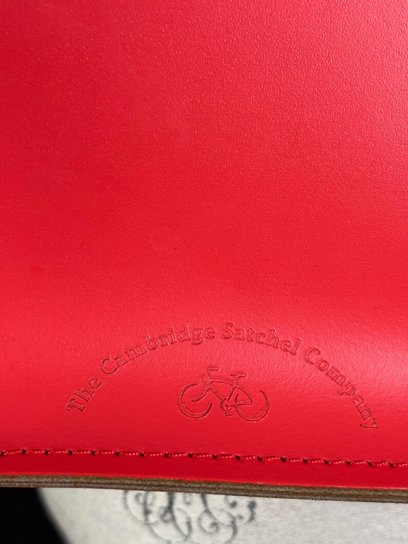 May include: Close-up of a bright red leather satchel. The Cambridge Satchel Company logo is embossed on the leather, featuring a bicycle icon. The leather has a textured appearance and a visible seam.