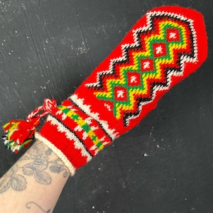 Può includere: Una muffola lavorata a maglia di colore rosso acceso con un motivo geometrico a rombi in giallo, verde, nero e bianco. Il polsino è rifinito con un bordo a coste bianco e una nappa decorativa in filato multicolore fissata con un cordino.