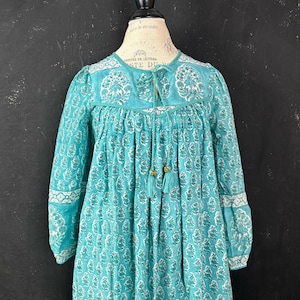 Puede incluir: Un vestido turquesa con un estampado floral blanco y mangas largas. El vestido presenta un cuello con cordón, borlas doradas y ribete blanco en las mangas y el escote. El vestido se exhibe en un maniquí.