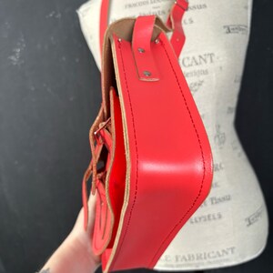 May include: A bright red leather satchel bag with a long adjustable strap. The bag features a flap closure with decorative stitching and a buckle detail. The interior is a lighter shade of brown. The bag is shown on a mannequin.