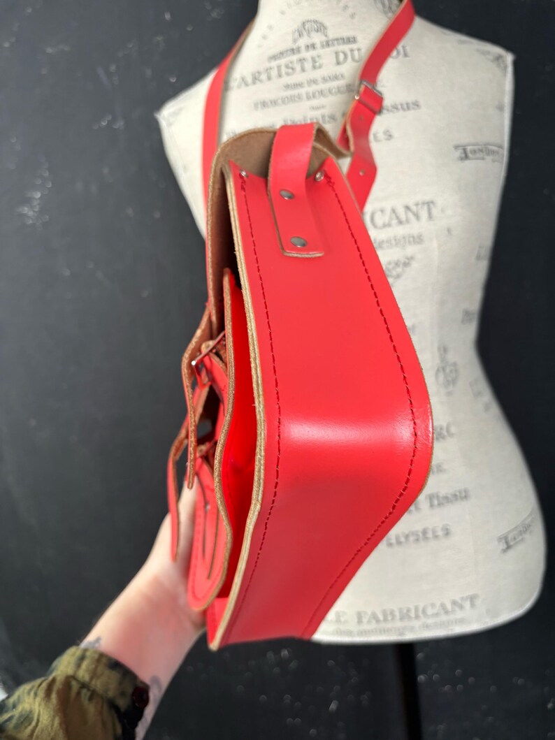 May include: A bright red leather satchel bag with a long adjustable strap. The bag features a flap closure with decorative stitching and a buckle detail. The interior is a lighter shade of brown. The bag is shown on a mannequin.