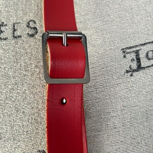 May include: Close-up of a bright red leather belt with a silver-colored buckle. The belt has a rectangular buckle and a single rivet. The background is a textured, light-colored fabric.