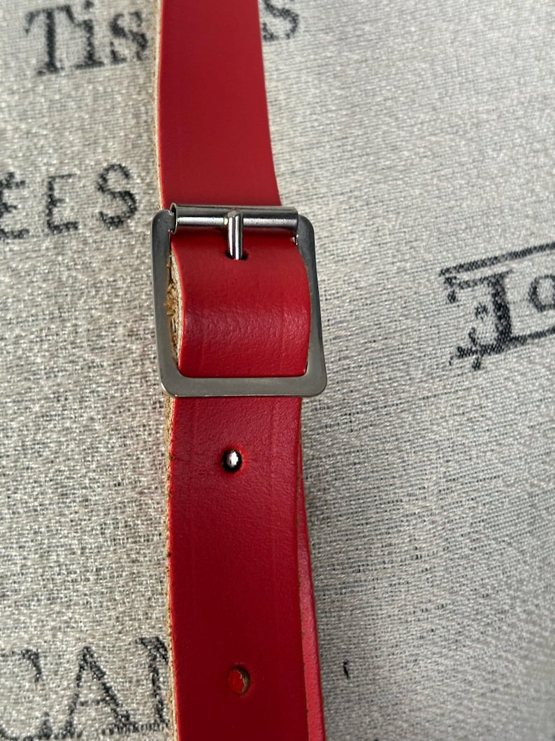 May include: Close-up of a bright red leather belt with a silver-colored buckle. The belt has a rectangular buckle and a single rivet. The background is a textured, light-colored fabric.