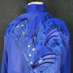 Può includere: Un maglione blu reale con collo alto. Il maglione presenta un design decorativo sulle spalle e sulla parte superiore della schiena, che incorpora varie tonalità di blu, paillettes e dettagli ritagliati. Il design ha un motivo floreale o a forma di foglia.
