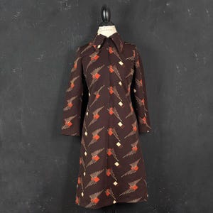 Pode incluir: Um vestido castanho escuro pelo joelho com fecho de correr frontal, gola pontiaguda e mangas compridas. O tecido é decorado com um padrão geométrico repetitivo de quadrados cor de laranja e linhas diagonais finas em tom creme.