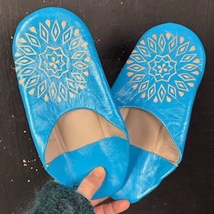 Puede incluir: Un par de zapatillas de cuero turquesa brillante. Cada zapatilla presenta un interior beige y un diseño geométrico dorado en la parte superior. Las zapatillas se muestran sobre una superficie oscura.