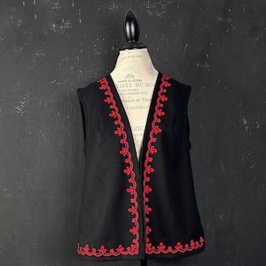 Puede incluir: Un chaleco negro sin mangas con un vibrante patrón floral bordado en rojo a lo largo de la abertura frontal y el dobladillo inferior. La prenda se exhibe en un maniquí, mostrando un escote en V profundo y un diseño abierto estructurado.