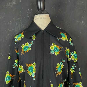 Pode incluir: Um casaco preto com fecho de correr e um padrão floral com flores turquesas, amarelas e cor de laranja. O casaco tem um painel central preto sólido, uma gola pontiaguda e mangas compridas. A peça é exibida num manequim de cor creme.