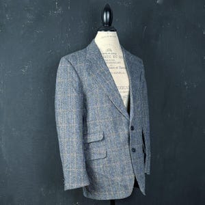 Può includere: Un blazer a quadri blu e grigio su un manichino. La giacca ha due tasche anteriori e due bottoni. Il tessuto ha un aspetto strutturato. Il manichino è posto su uno sfondo scuro.