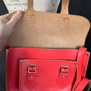 May include: A red leather satchel with a flap closure and a front pocket. The bag has a long adjustable strap and silver-colored buckles. The interior is tan-colored. The bag is open, revealing the inside. The text "ARTISTE DU I" is visible.