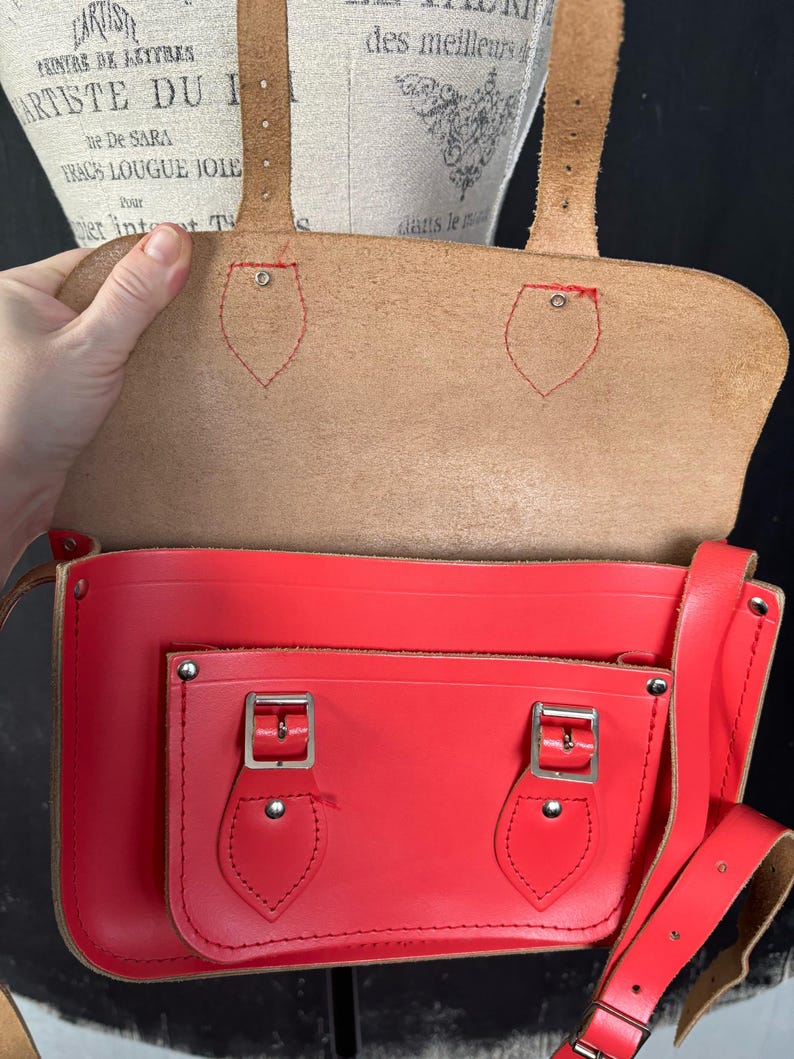 May include: A red leather satchel with a flap closure and a front pocket. The bag has a long adjustable strap and silver-colored buckles. The interior is tan-colored. The bag is open, revealing the inside. The text "ARTISTE DU I" is visible.