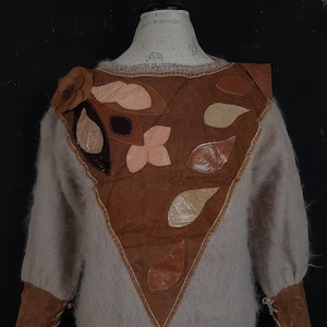 Peut inclure: Un pull beige avec un motif patchwork en cuir marron et peau de serpent sur le devant. Le pull présente un accent floral et des poignets en cuir avec des détails de laçage. Le design est présenté sur un mannequin.