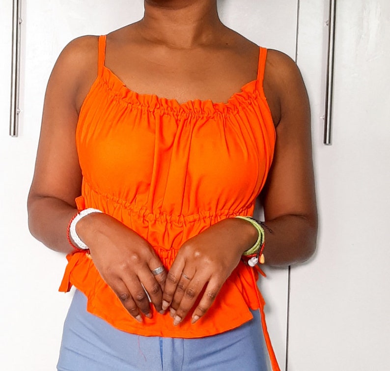 May include: A bright orange, sleeveless top with a ruffled neckline and a cinched waist. The top has adjustable straps and a tie at the back.