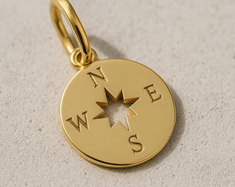 May include: A gold-colored compass pendant with a loop at the top. The compass rose is cut out in the center of the pendant. The cardinal directions N, E, S, and W are engraved around the compass rose.
