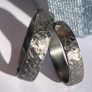 Könnte beinhalten: Zwei strukturierte Silberringe mit einem geometrischen Muster. Die Ringe werden auf einer weißen Oberfläche mit einem Schatten und einer blauen Samtringbox im Hintergrund präsentiert. Die Ringe scheinen Eheringe zu sein.