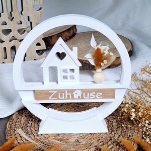 Könnte beinhalten: Weiße Holz-Deko mit einem kleinen Haus-Ausschnitt, einem Herz und dem Wort "Zuhause" auf einer Holztafel. Die Deko steht auf einem weißen Sockel und hat eine kleine Vase mit Trockenblumen.