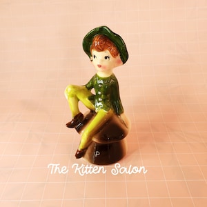 May include: A ceramic figurine of a seated boy in a green outfit and hat, with brown shoes. The boy is sitting on a brown mushroom. The figurine has a painted face with rosy cheeks. The text "The Kitten Salon" is at the bottom.