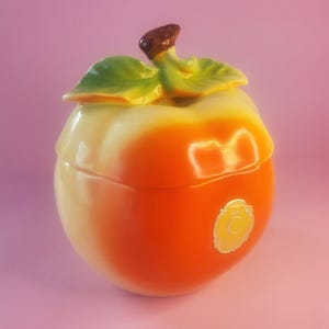 May include: A ceramic apple-shaped jar with a lid, featuring a gradient of colours from yellow to orange. The jar has green leaves and a brown stem on top. A gold-coloured emblem is on the side.