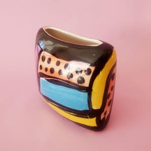 May include: A small, rounded ceramic vase with a glossy finish. The surface features a bold, abstract design with thick black outlines, a bright blue rectangular centre, a peach-coloured section with black dots, and vibrant yellow accents.