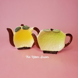 May include: Two decorative ceramic teapots. One is yellow with brown accents and green leaf details. The other is light green with a cracked texture and brown accents. The Kitten Salon text is visible.