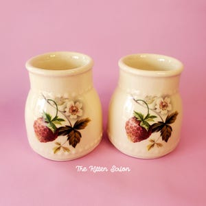 May include: A pair of cream-coloured ceramic jars, each decorated with a strawberry motif. The design includes a red strawberry, green leaves, and white flowers. The jars have a flared rim and a rounded shape, set against a pink backdrop.