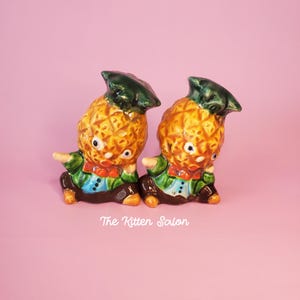 May include: Two vintage ceramic pineapple figurines. Each figurine has a yellow and brown pineapple body, green hat, and a green jacket with a red bow tie. The figurines are on a pink background.