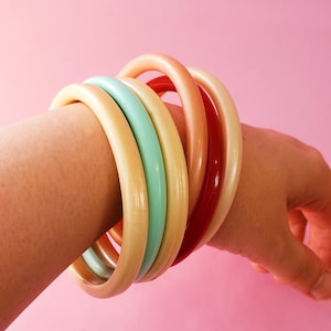May include: A selection of colourful bangles in cream, mint green, coral, and red. The bracelets are stacked on a wrist, showing their smooth, rounded design. The background is a solid pink.