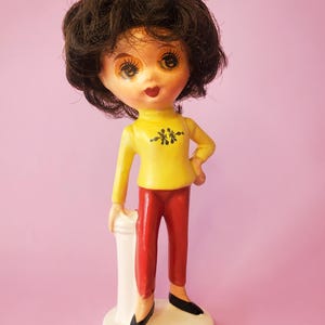 May include: A vintage ceramic doll with dark hair, wearing a yellow top with black floral accents and red trousers. The doll stands on a white base with a blue trim, and black shoes. The doll is posed with one hand on a white pillar.