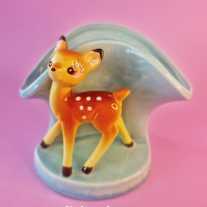 May include: A ceramic figurine of a fawn with brown fur, white spots, and a yellow face, standing in front of a light blue, curved, shell-shaped holder. The text "The Kitten Salon" is printed in white at the bottom of the object.