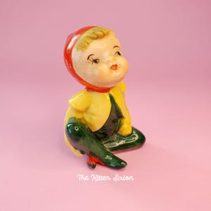 May include: A vintage ceramic figurine of a seated child. The figurine has a red hood, yellow jacket, and green trousers. The child's face is painted with rosy cheeks and a small mouth. The Kitten Salon is printed on the bottom.