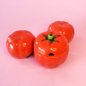 May include: Three ceramic tomato-shaped kitchen items. Two are salt and pepper shakers with small holes on top. The third is a lidded container with a green stem and a small opening. All are glossy red with green and black accents.