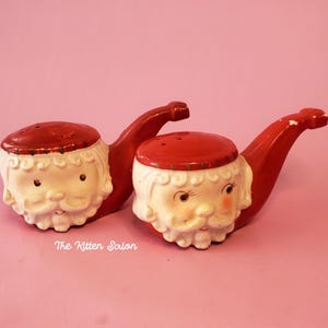 May include: A pair of vintage Santa Claus salt and pepper shakers. Each has a red hat and pipe, with a white beard and face. The text "The Kitten Salon" is visible on the bottom.