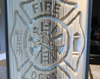 Può includere: Un libro piegato in una scultura tridimensionale di un logo del dipartimento dei vigili del fuoco. Il logo è una croce bianca con le parole "Fire Department" all'interno.