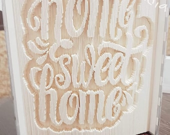 Può includere: Un libro bianco piegato in una scultura 3D delle parole "home sweet home". La scultura è su una superficie di legno.