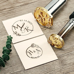 Könnte beinhalten: Zwei Holzbrandwerkzeuge mit goldfarbenen Stempeln und Holzquadraten. Die Stempel zeigen Initialen und Daten, darunter "M&K 02.14.2025" und "H&A 02.14.25", umgeben von floralen Designs. Die Werkzeuge haben silberne und schwarze Griffe.