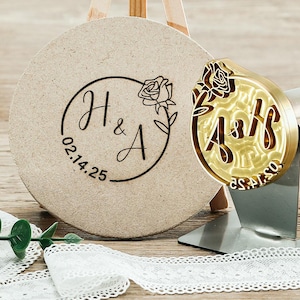 May include: A custom wedding stamp with the initials "H & A" and the date "02.14.25" in a circular design with a rose. The stamp is made of gold metal and is next to a round, beige coaster with the same design.